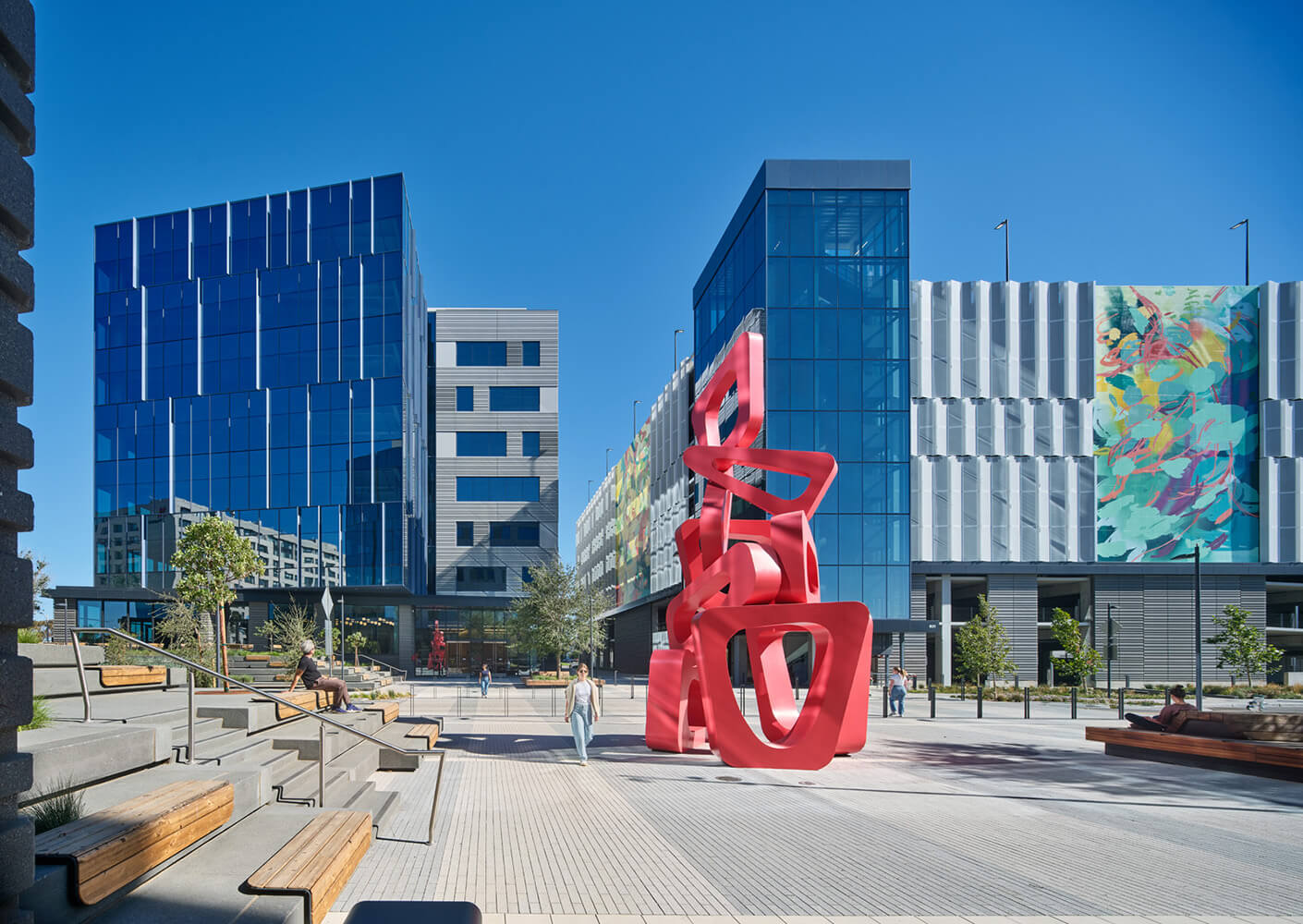 Modern plaza with red sculpture, blue glass buildings, and seating area on a sunny day.