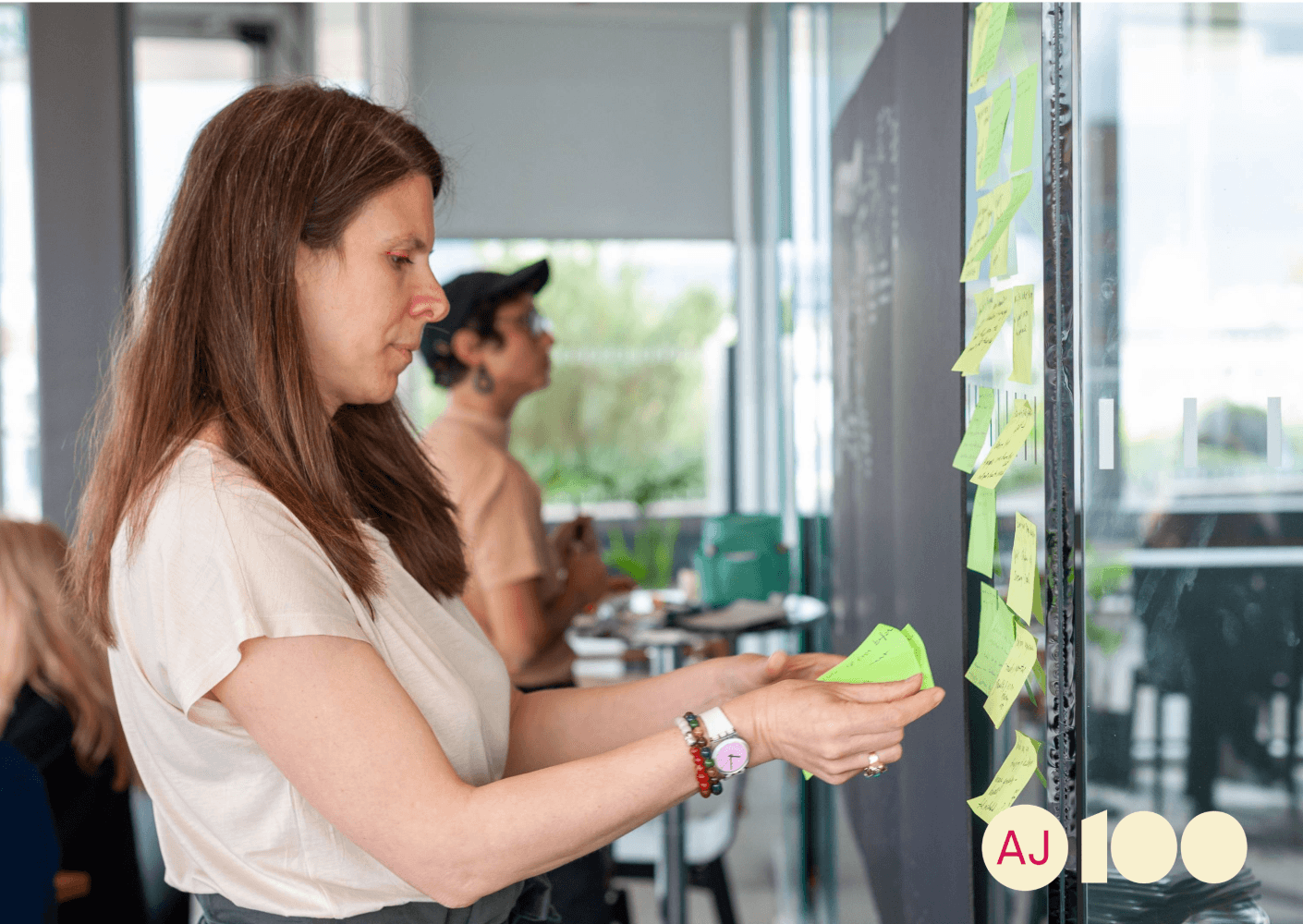 Woman placing sticky notes on a glass board during a brainstorming session. Another participant is visible in the background.