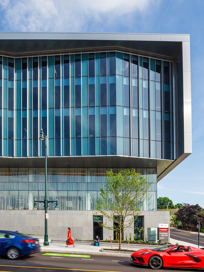 Modern glass hospital building with pedestrians and cars on a sunny day.