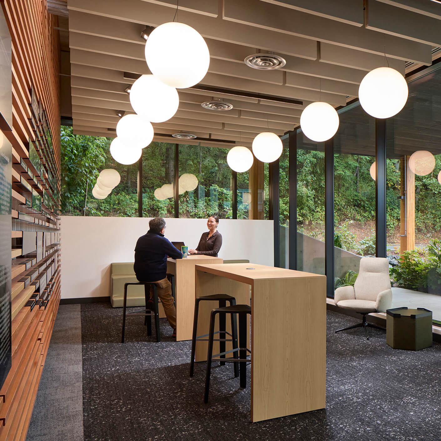 Modern office lounge with two people meeting, bright lighting, and large windows overlooking greenery.
