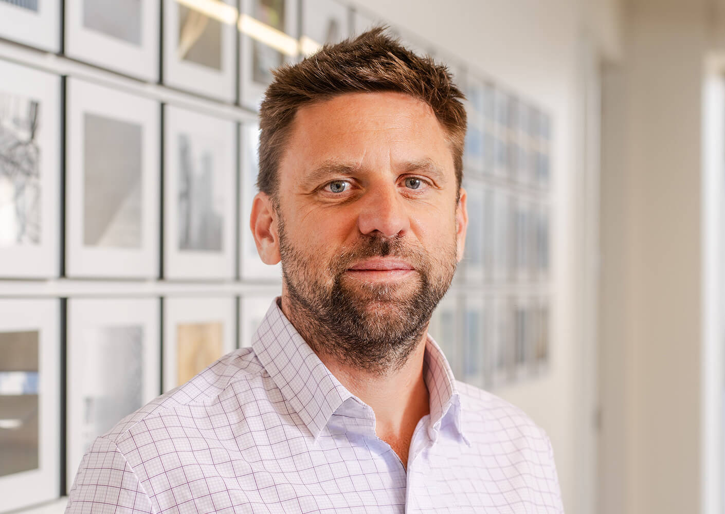 Portrait of a man with a beard in a checkered shirt, standing in front of a gallery wall featuring framed images.