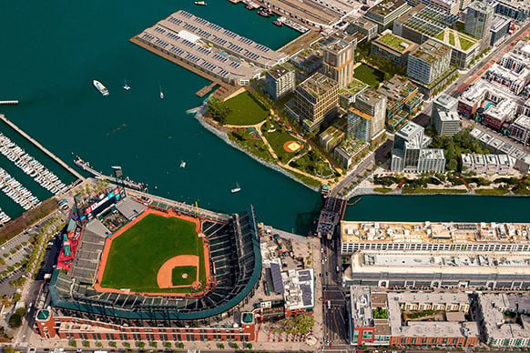 Aerial view of a waterfront area featuring a baseball stadium, residential buildings, a marina, and landscaped parks along the water's edge.