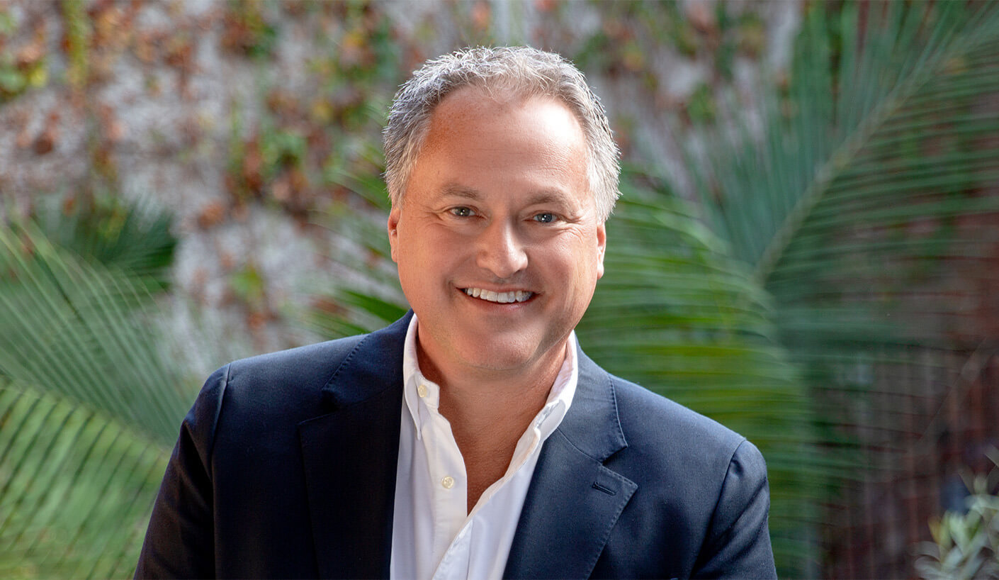 Headshot of a smiling man in a dark blazer and white shirt, surrounded by green plants, against a textured background.