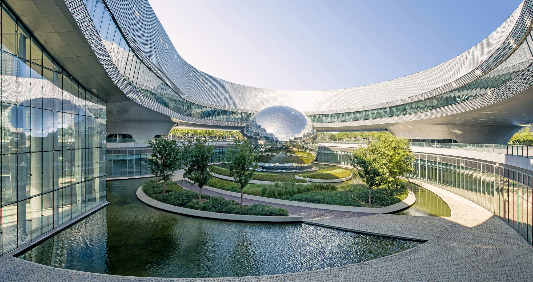 A unique architectural structure featuring a reflective sphere, surrounded by greenery and water elements, under a clear blue sky.