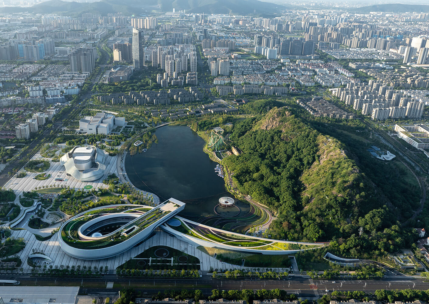 Aerial view of a city park featuring a lake, landscaped gardens, and contemporary buildings set against a backdrop of urban development and hills.