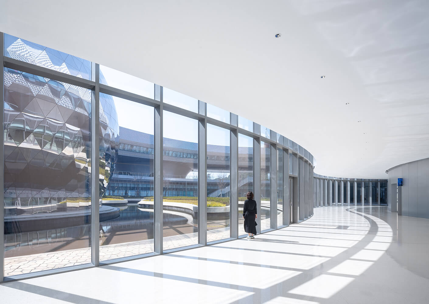 Interior view showcasing large windows and a curved corridor, with a figure walking alongside a reflecting pond and landscaped area.