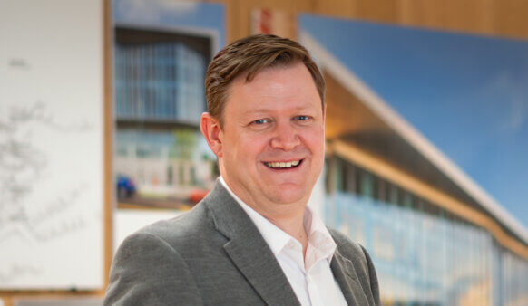 Portrait of a smiling professional man in a suit, standing in an office with architectural designs in the background.