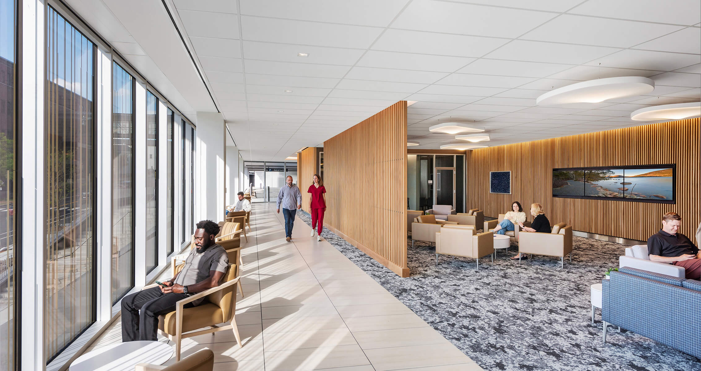 Modern hospital lobby with people sitting and walking, large windows, and wooden accents.