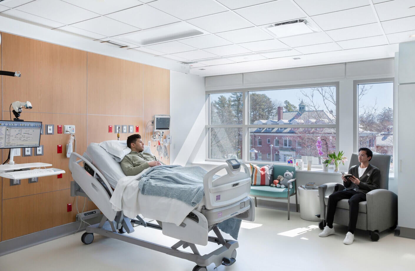 Patient resting in hospital room with visitor by window.