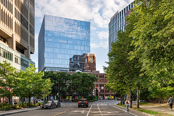 City street scene featuring glass office buildings and historic clock tower, with trees lining the road and vehicles and bicycles visible.
