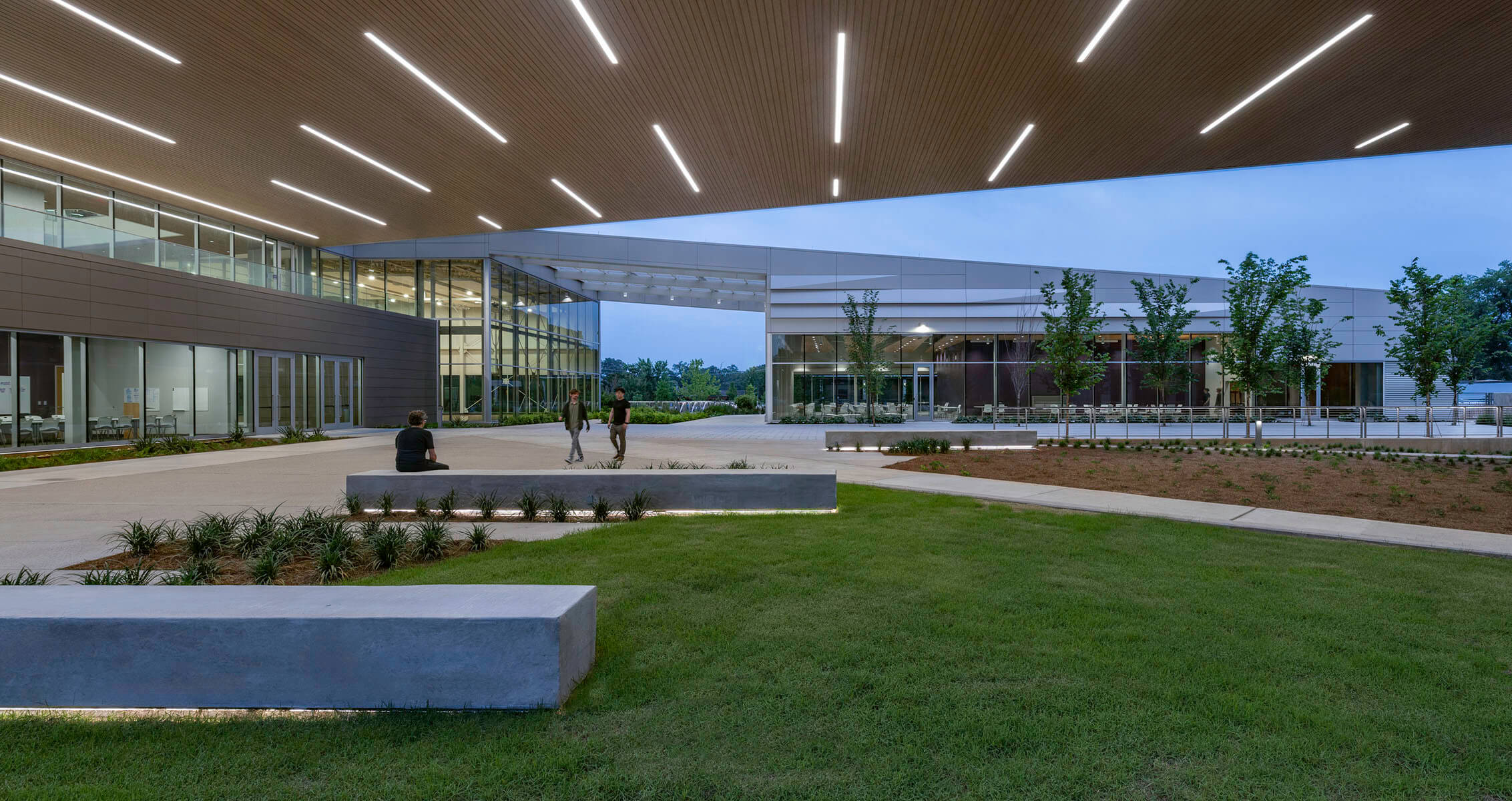 Modern building exterior with people walking, green lawn, and illuminated ceiling panels.