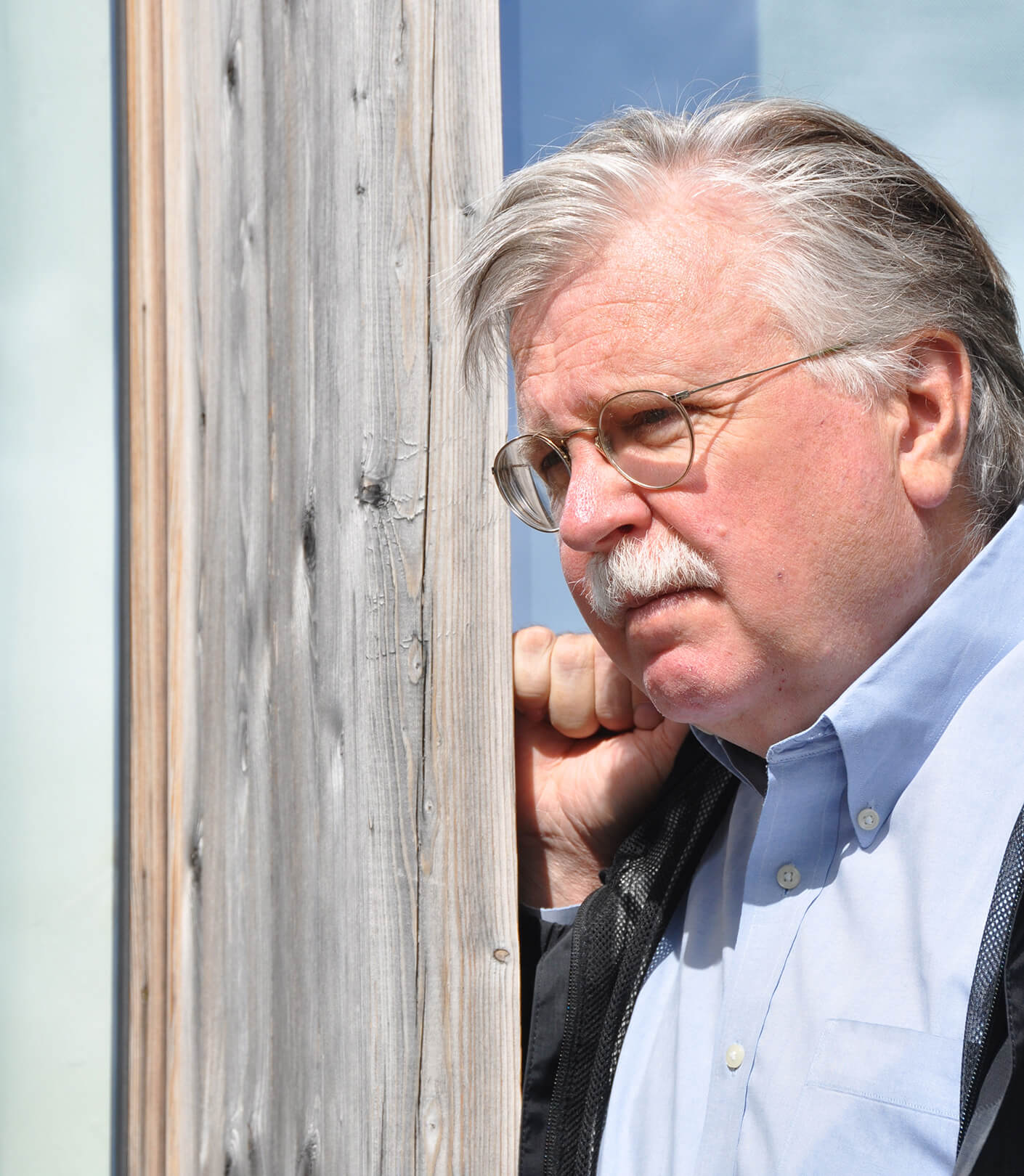 Left profile of Ralph Johnson leaning against a pale gray wooden doorframe in blue oxford and sport jacket.