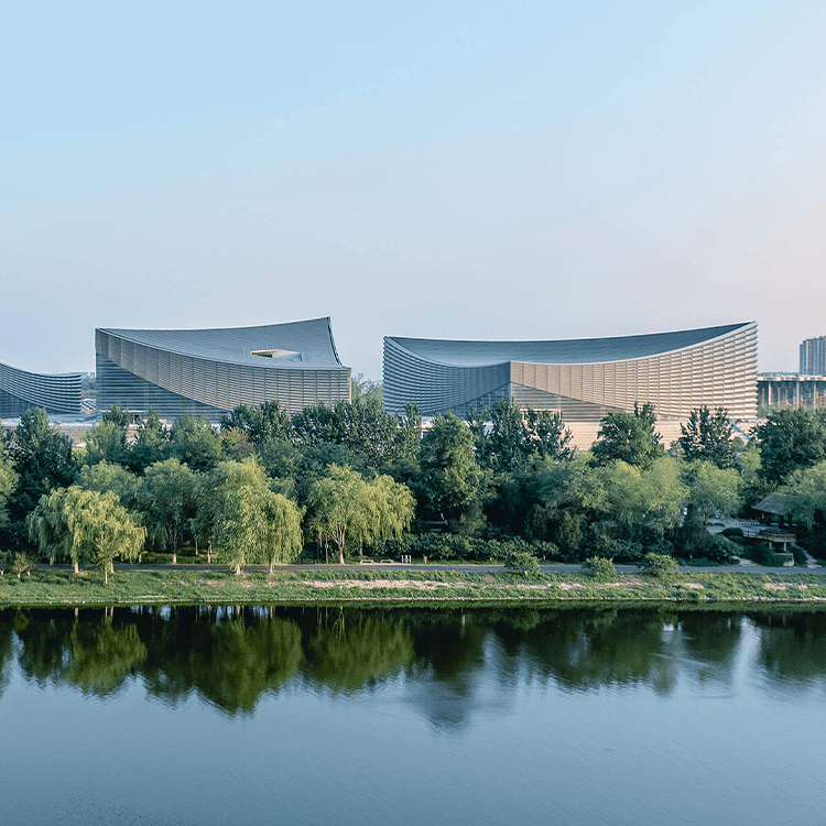 Architectural view of unique buildings beside a waterway, surrounded by greenery and trees, reflecting in the water.