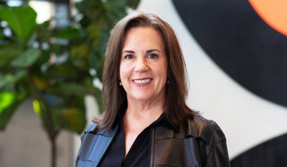 Professional woman smiling in front of a colorful backdrop with plants, wearing a black shirt and leather jacket.