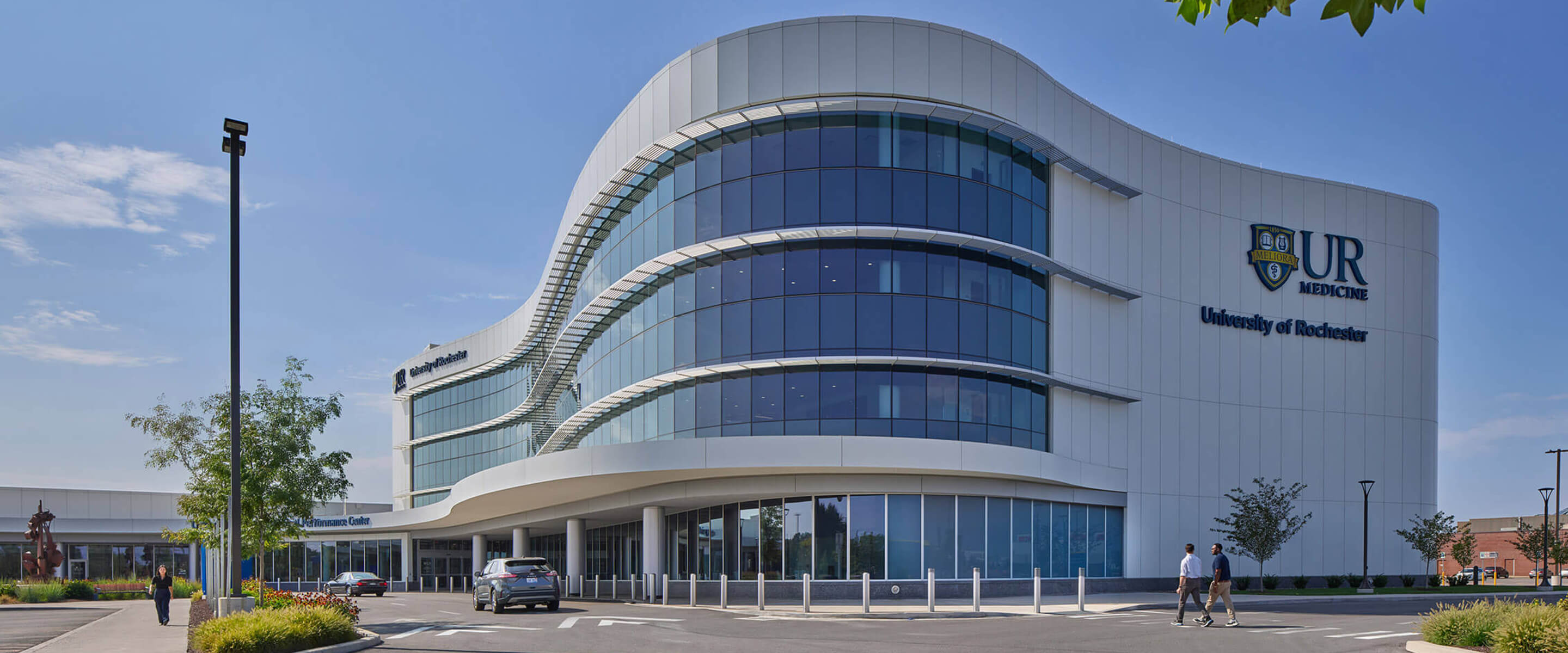 Image of the University of Rochester Medical Center featuring a sleek glass facade and signage. Visitors walking past the entrance on a sunny day.