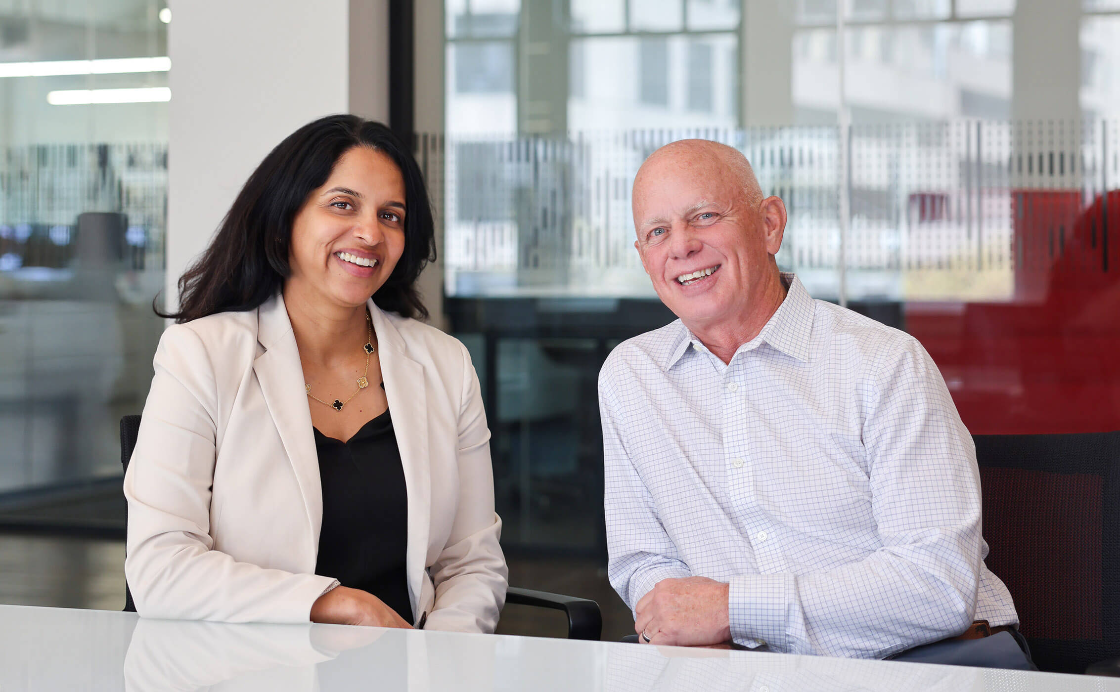 Vandana and Tom sit side by side in a bright conference room, smiling