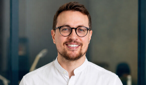 Smiling man with glasses wearing a white shirt, posing in an office setting with a neutral background.