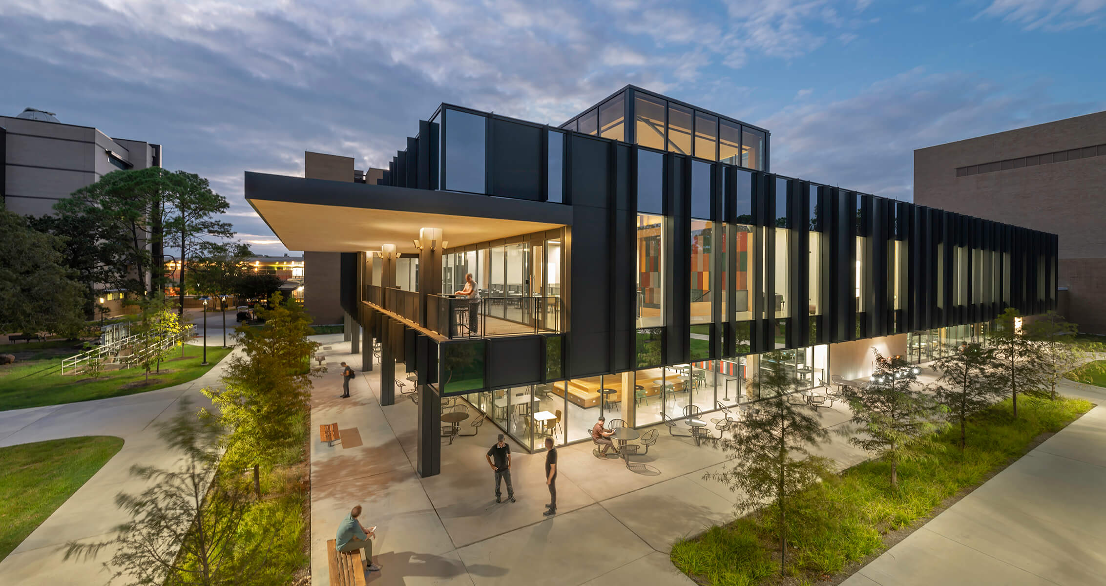 Modern glass building exterior with people outside at dusk, surrounded by trees and pathways.