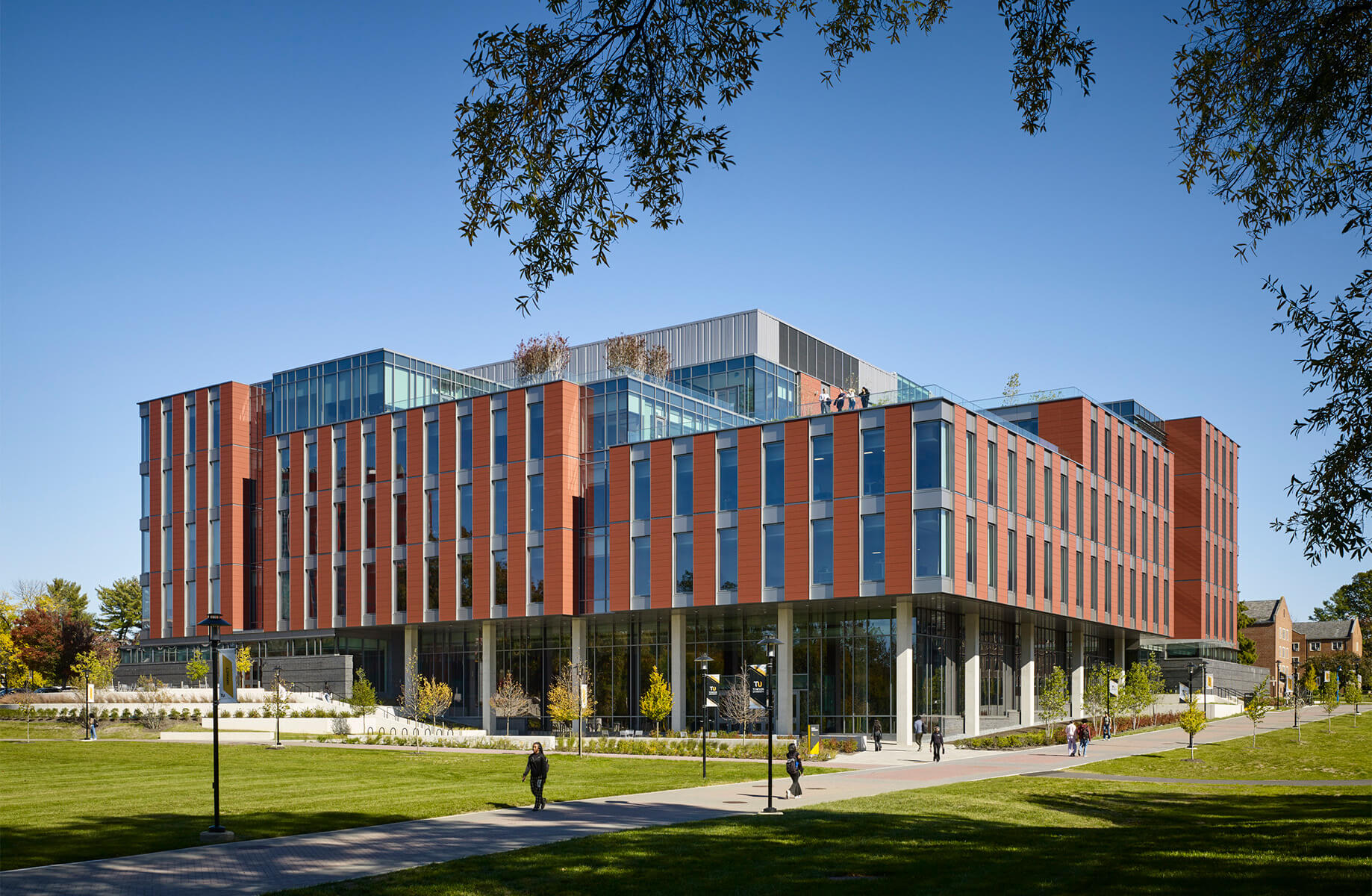 Modern university building with red brick facade and glass windows on a sunny day, surrounded by green lawn.