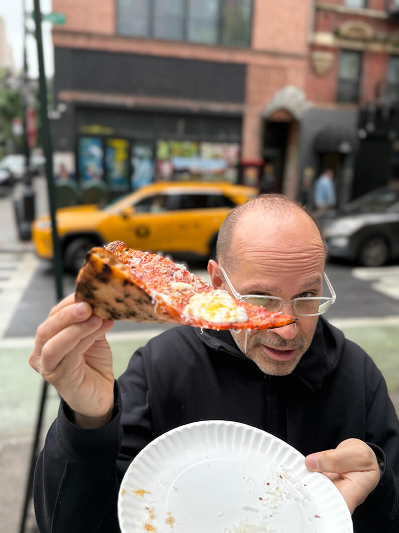 Man holding a pizza slice on a street with a taxi in the background.