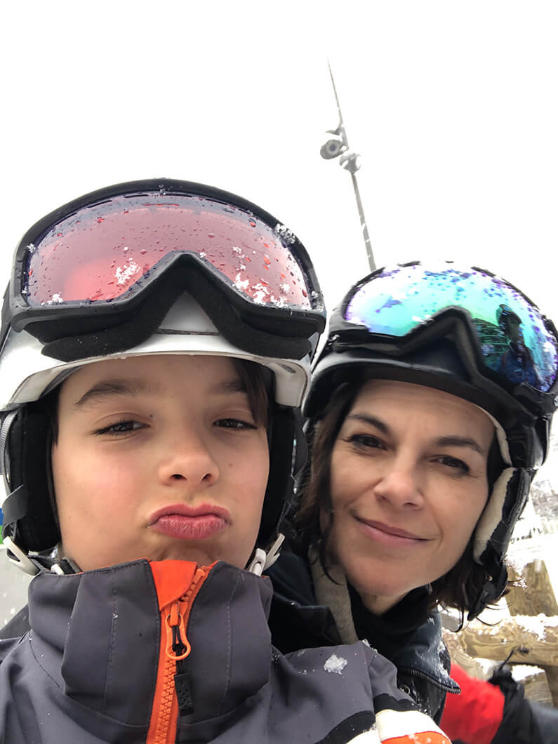 Mother and child on a ski trip, both wearing helmets and goggles, with snowy background.