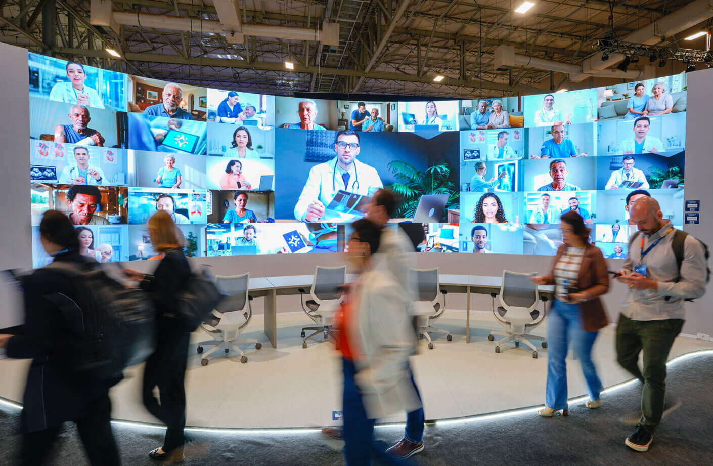 Conference attendees pass large video wall displaying virtual meeting participants.