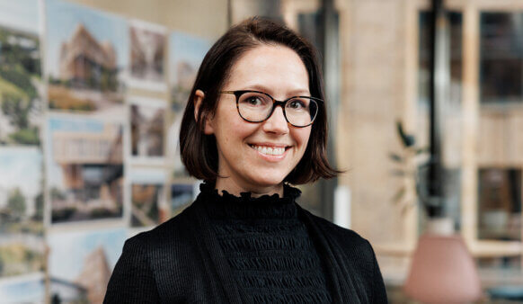 Smiling woman with glasses in office setting, blurred design boards in background.