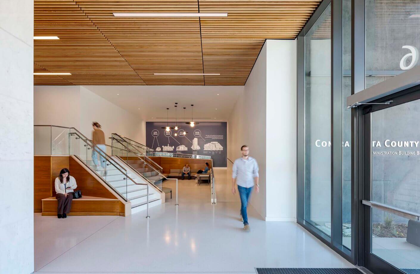 Modern office lobby with wood ceiling, staircase, and people walking and sitting.