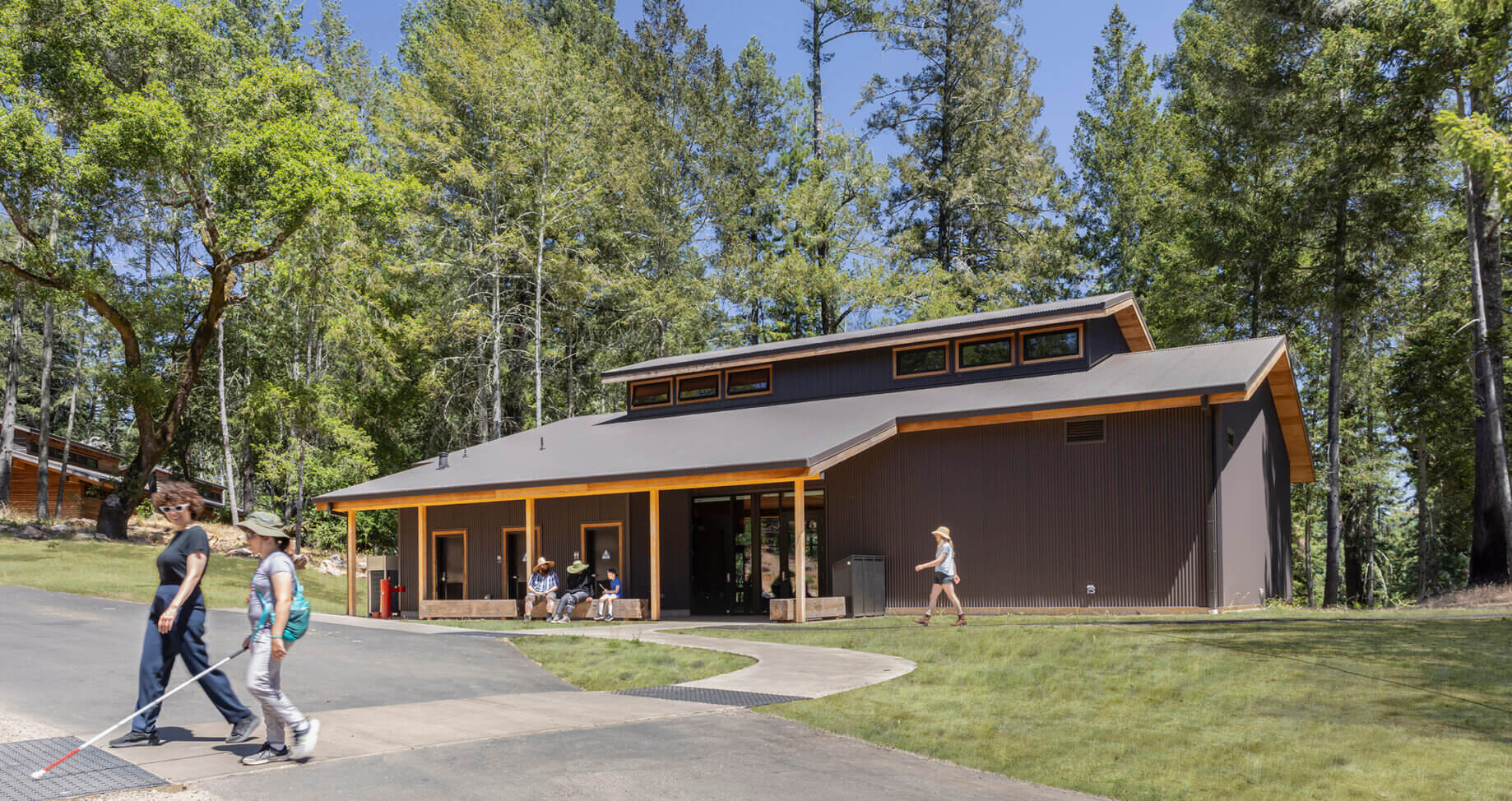 Two people walk past a shaded building surrounded by trees, with additional visitors sitting near the entrance on a sunny day.