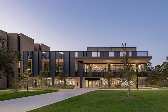 Modern building exterior with glass facade at dusk, surrounded by greenery.