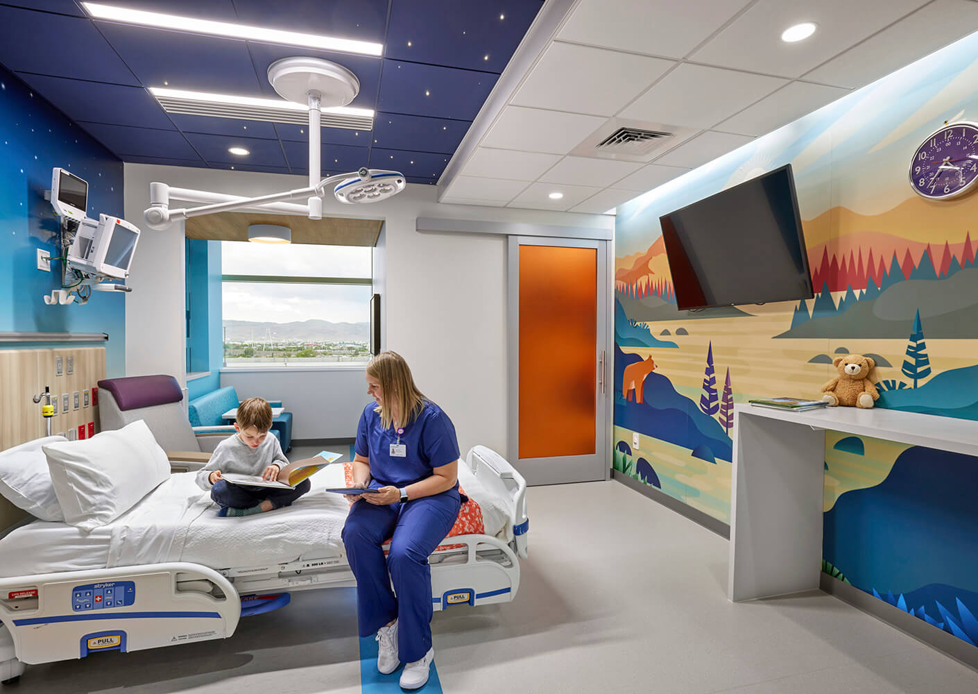 Pediatric patient room with twinkle lights in the ceiling and nature mural on the wall, plus nurse and child reading on bed.