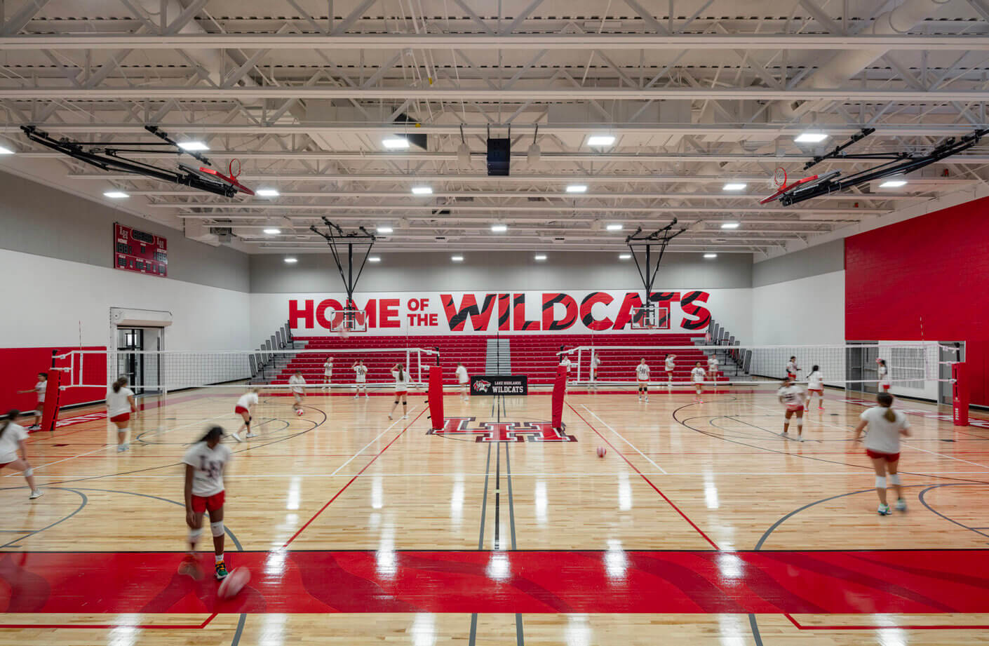 High school gym with bright red accents, students playing volleyball, Home of the Wildcats printed on the wall in background.