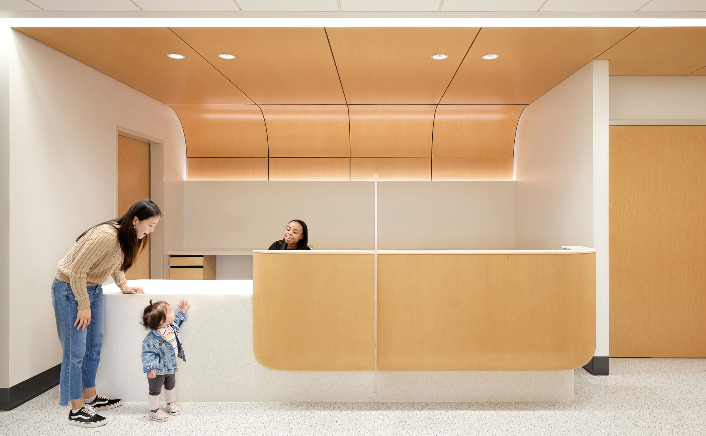 Modern reception area with a woman and child at a counter, sleek wood design.