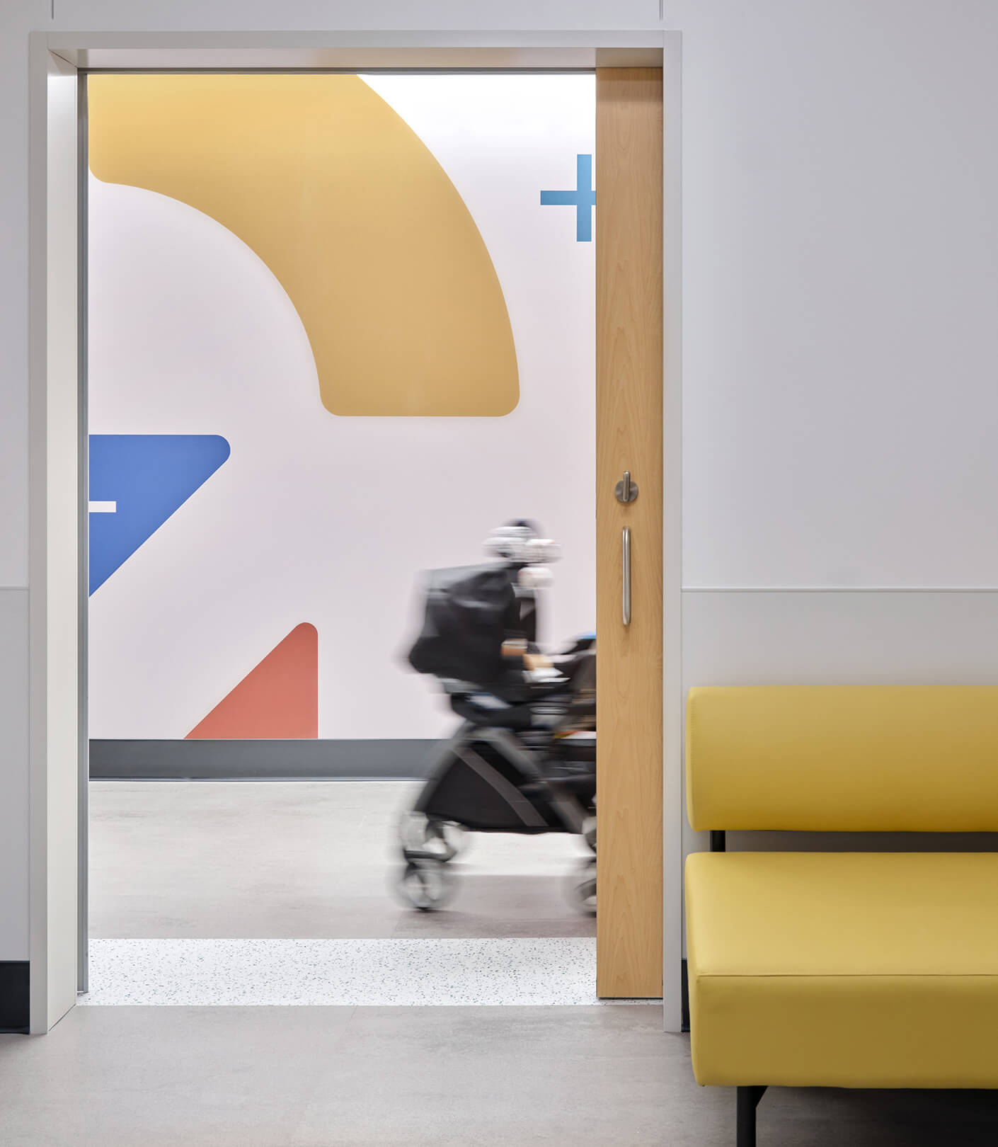 A blurred stroller passes outside a door open to a hallway lined with colorful wall design; inside, yellow seat in modern interior.