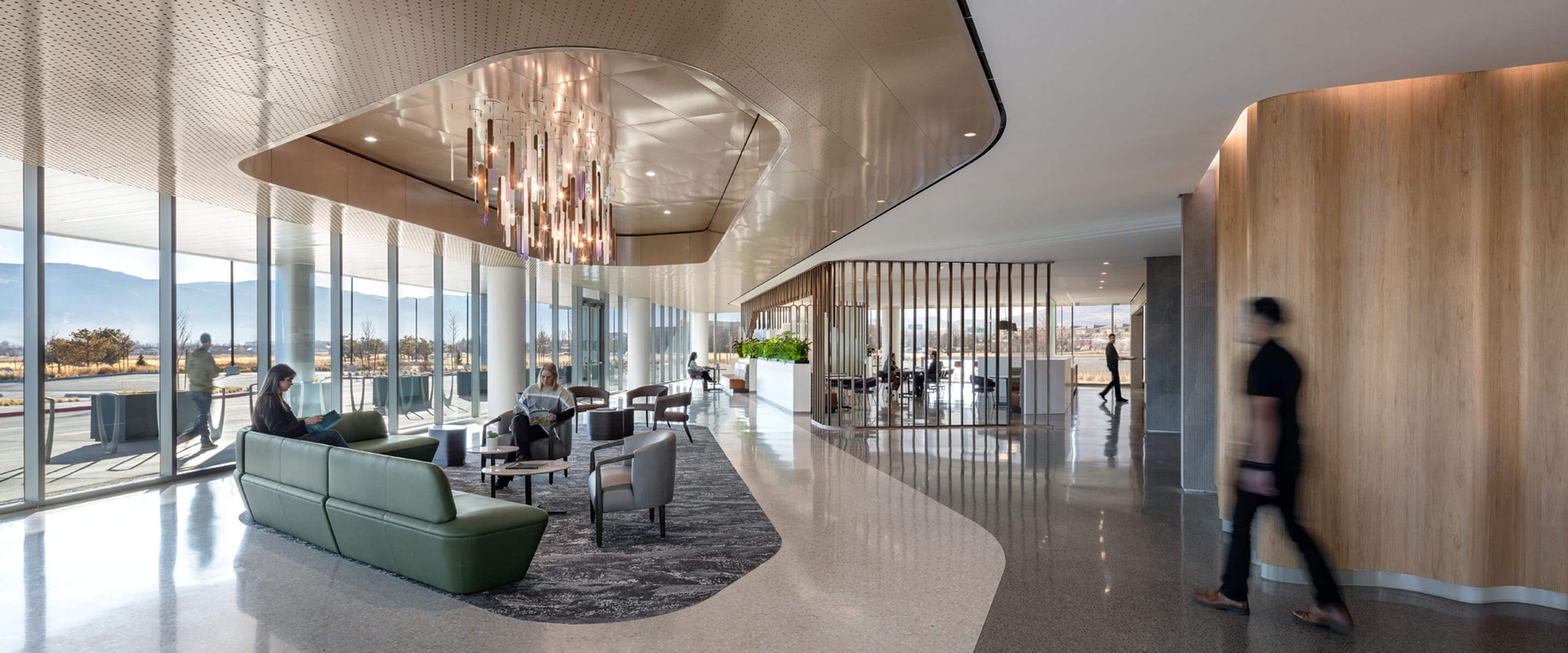 Lobby with seating under a chandelier and people walking along a curved path next to an undulating wood wall and floor-to-ceiling windows.