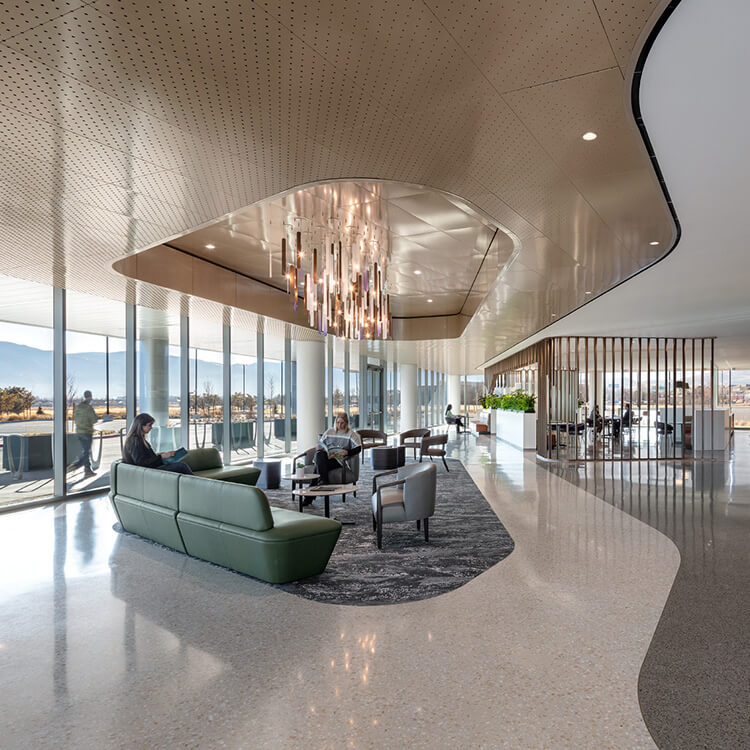 Lobby with seating under a chandelier and people walking along a curved path next to an undulating wood wall and floor-to-ceiling windows.