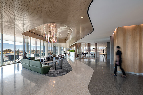 Lobby with seating under a chandelier and people walking along a curved path next to an undulating wood wall and floor-to-ceiling windows.
