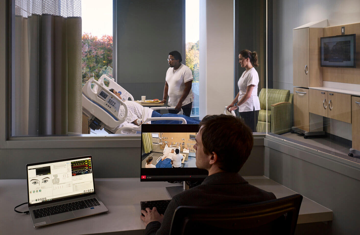 Healthcare simulation room with staff and monitoring equipment.