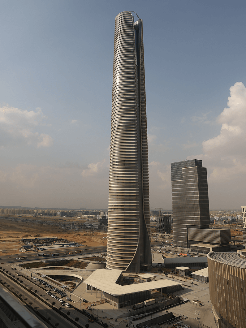 Tall modern skyscraper under blue sky with surrounding buildings; urban skyline scene.