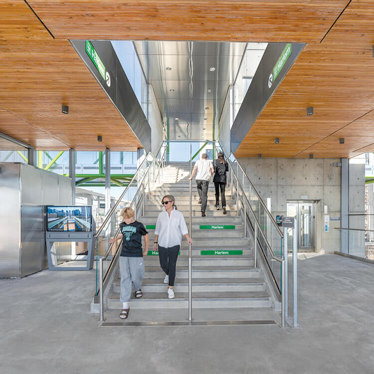 Passengers descend the stairs of a train station interior with a wood ceiling and green wayfinding signage.