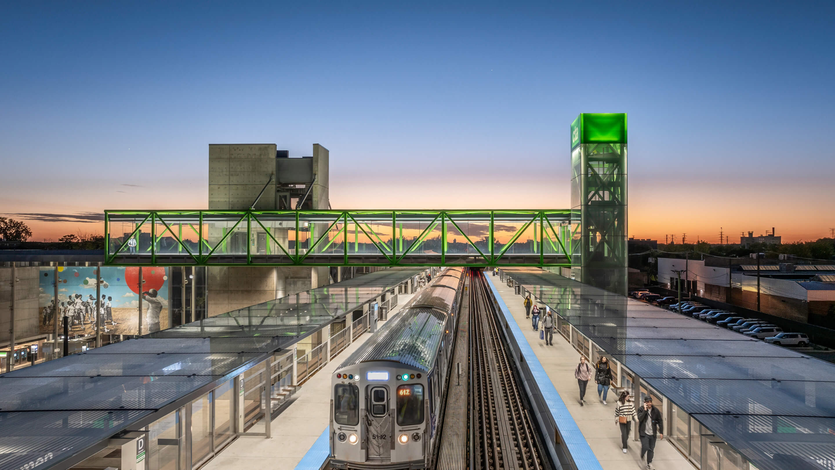 Modern train station at sunset with a green pedestrian bridge and a mural.