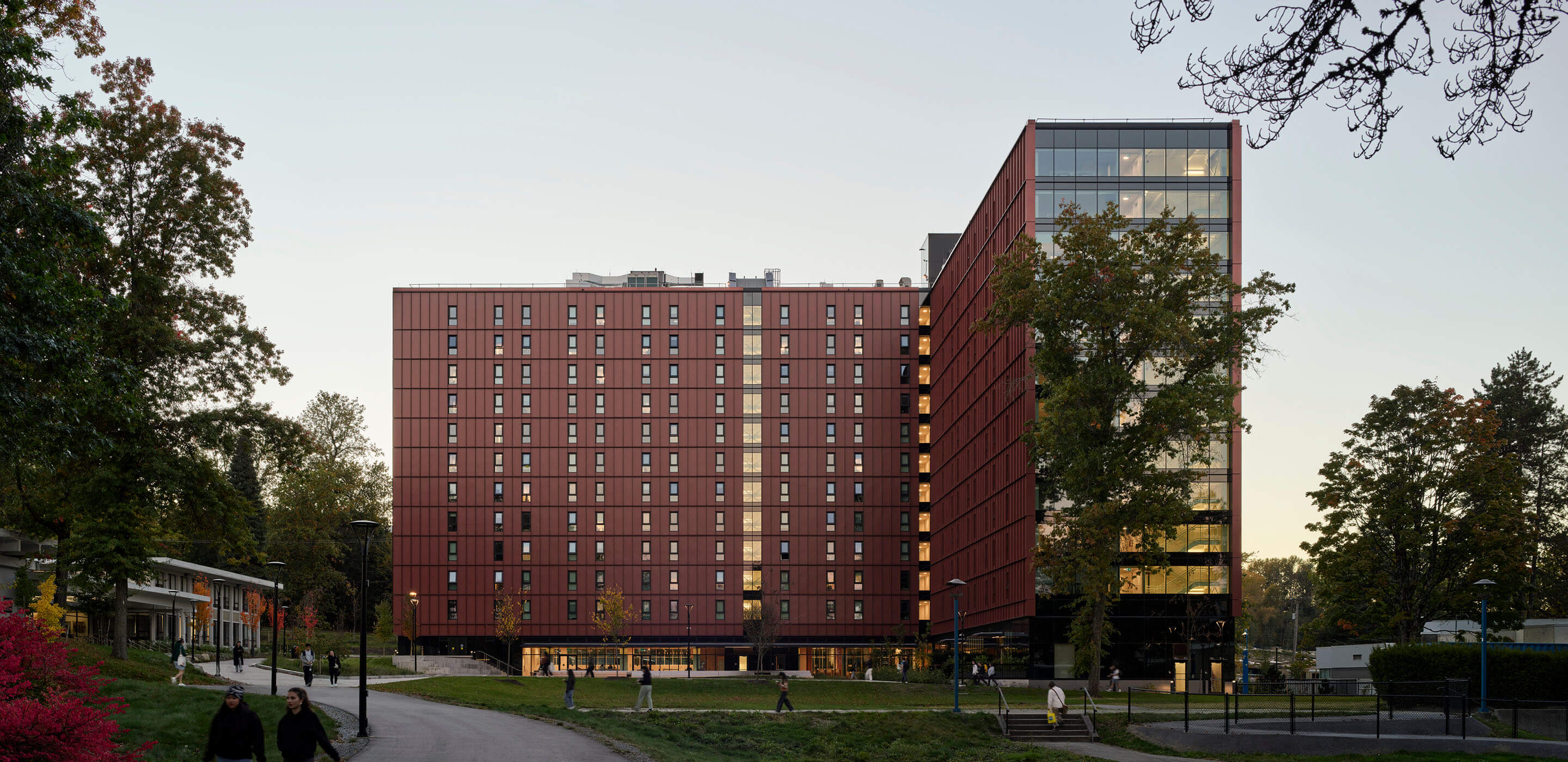 Modern red and glass building surrounded by trees, pathway, and people walking.