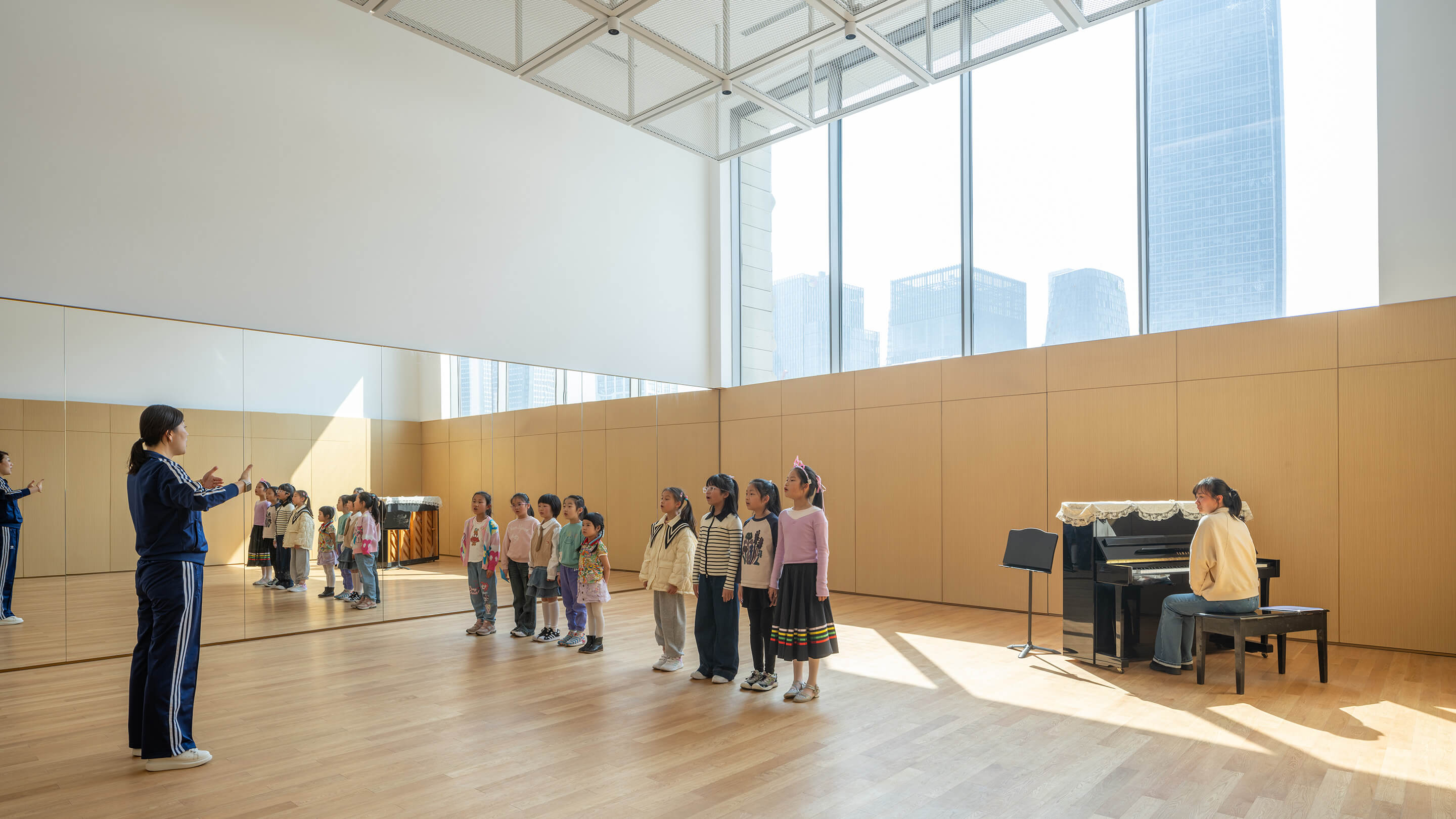 Children’s choir practice in bright music room with piano and instructor, city view outside.