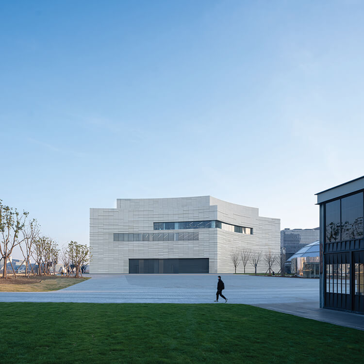Modern white building with curves and angles, set behind a paved plaza and grassy area on a winter day.