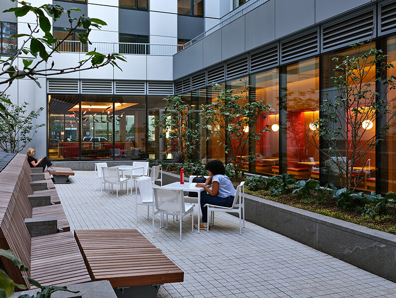 Outdoor terrace area with seating, plants, and people relaxing near a building.