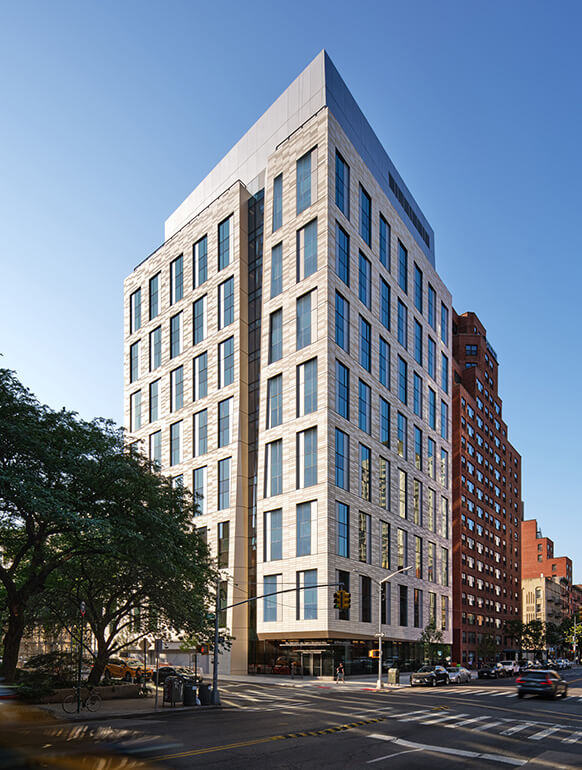 New building with large windows and stone facade at a city intersection, surrounded by trees and urban architecture. Clear blue sky above.