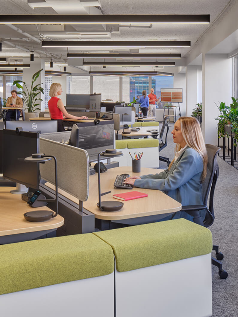 Office workspace featuring employees at desks with computers, plants, and collaborative areas. Bright lighting and ergonomic furniture enhance productivity.