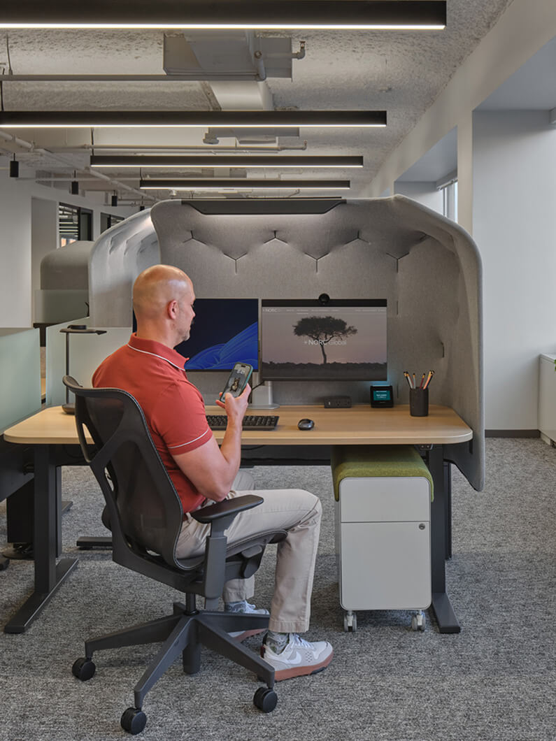Employee seated at a workspace in a cubicle, using a smartphone. Two monitors and office supplies are visible on the desk. Grey acoustic panels surround the area.