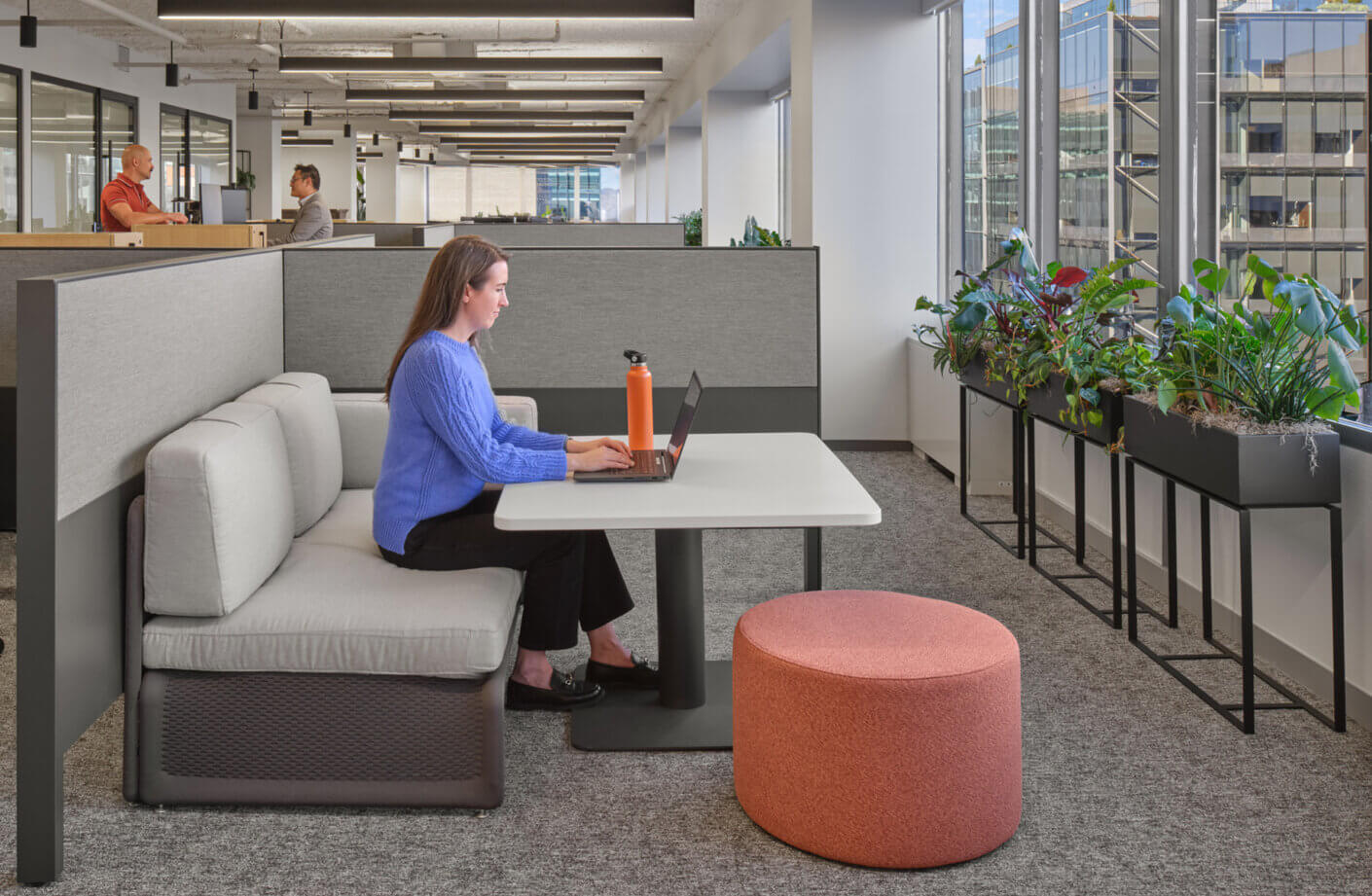 A woman working on a laptop at a collaborative workspace, featuring soft seating and plants along a large window. Two colleagues are visible in the background.