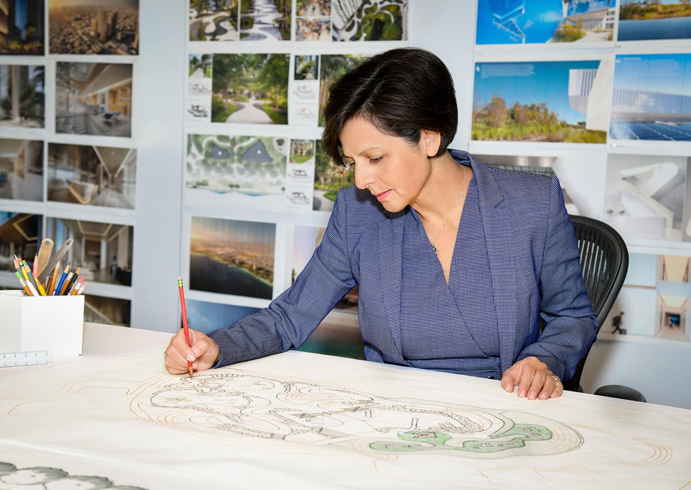 Female architect sketching a design plan surrounded by project visuals in an office.